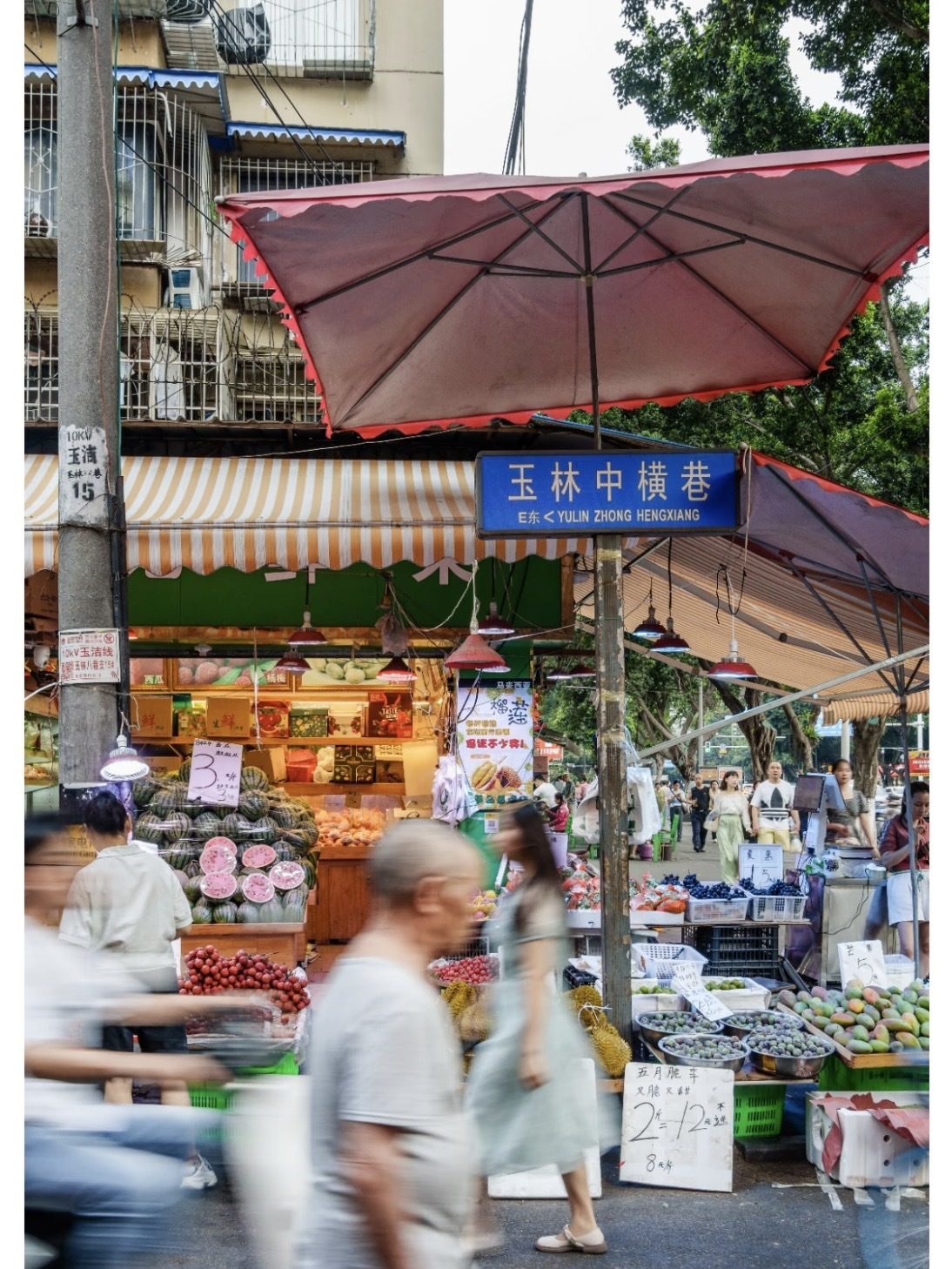 A bustling Chengdu street market in the Yulin neighbourhood, with fruit stalls under red umbrellas and locals shopping