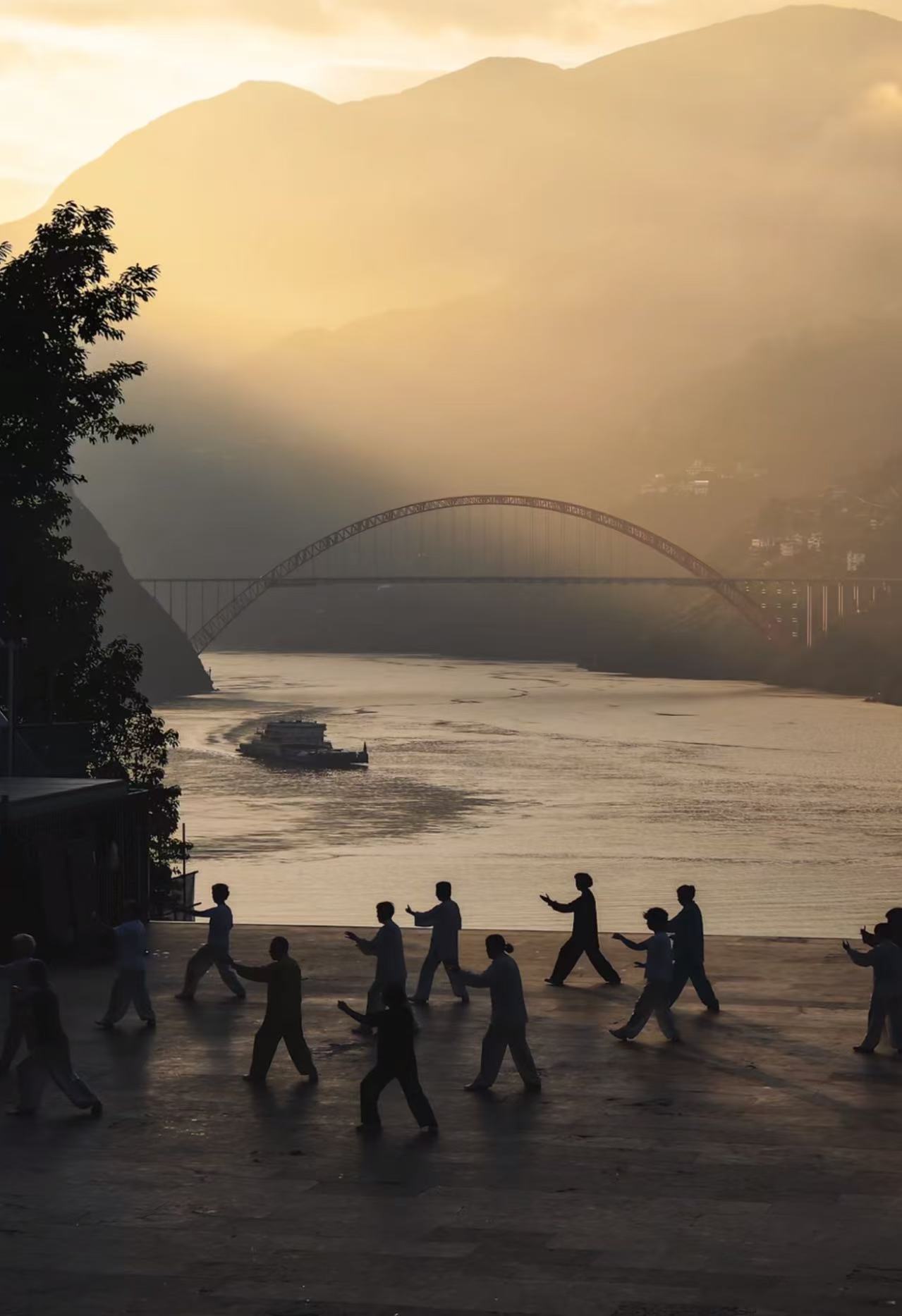 A group practicing Tai Chi at sunrise beside a river, with a bridge in the distance