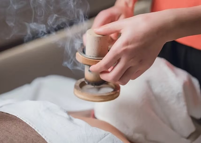 Close-up of a moxibustion treatment, with smoke rising from a heated tool during a traditional wellness session